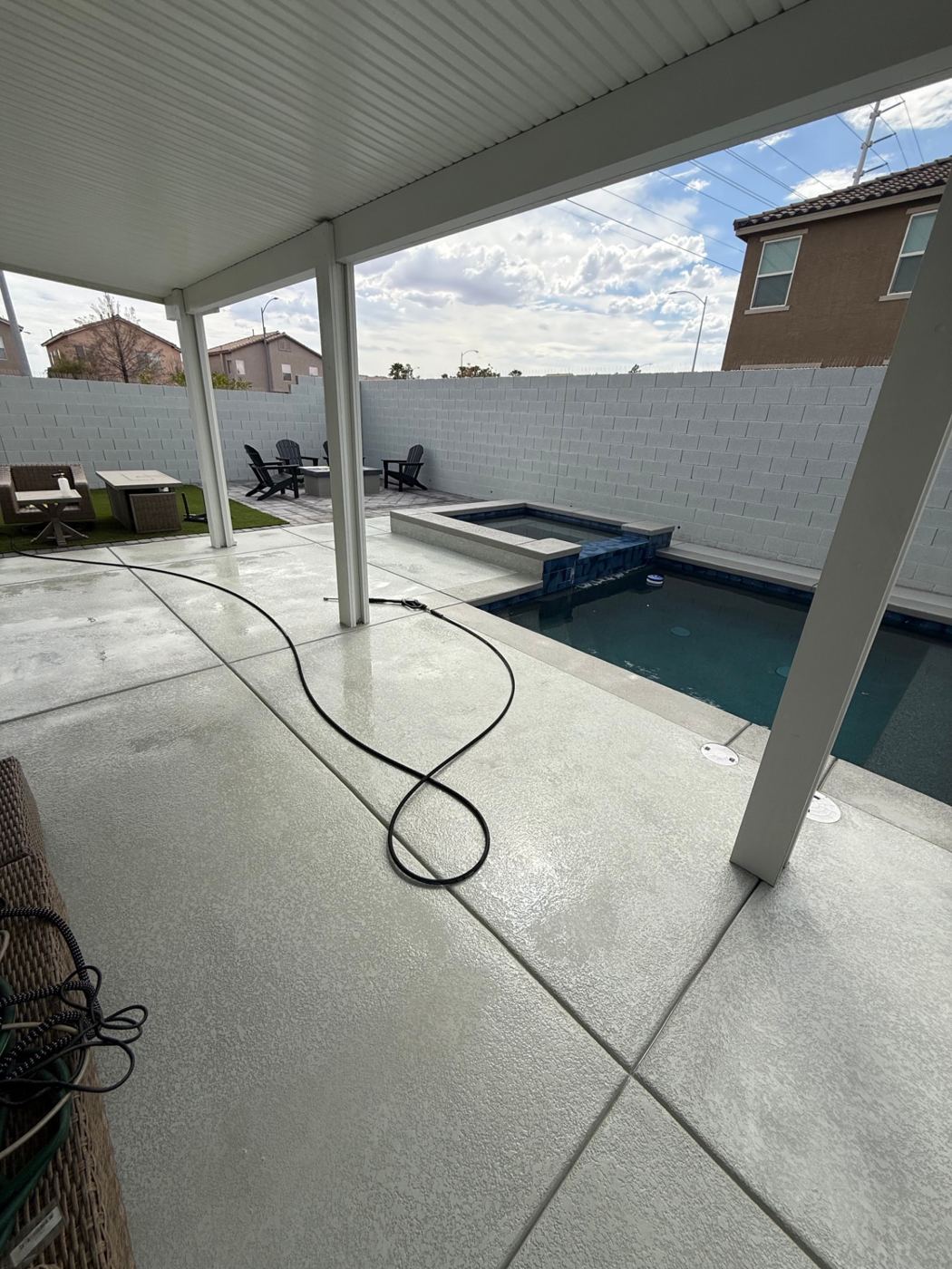 Covered backyard patio beside a pool, with a pressure-washing hose laid across the wet concrete deck